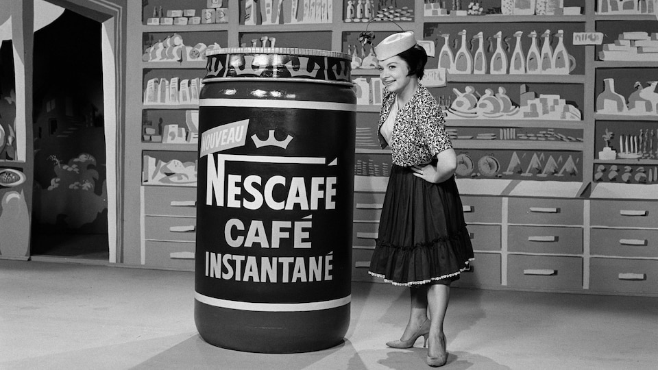 Mirielle Lachance dans un studio au décor d'épicerie à côté de contenant format géant de Nescafé.