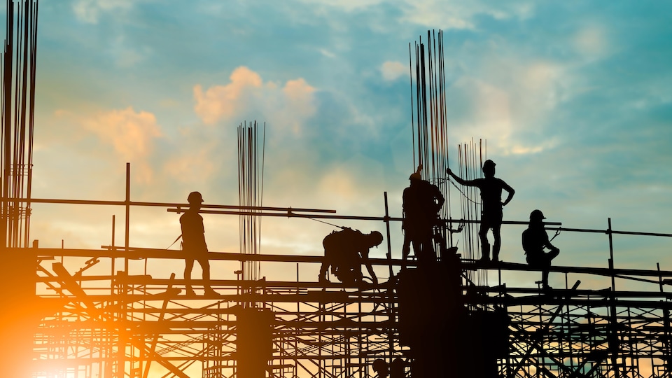 Les silhouettes de personnes sur une partie surélevée d'un chantier de construction sont vues lors d'un coucher ou lever du soleil. Ces personnes portent des casques protecteurs.