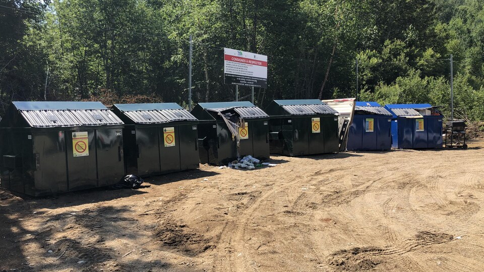 Six gros conteneurs à déchets sont alignés au fond d'un terrain sablonneux.