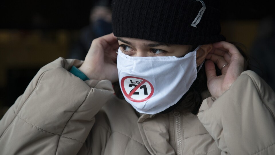 Une femme avec un masque noir sur lequel apparaît un écusson qui rejette la loi 21.