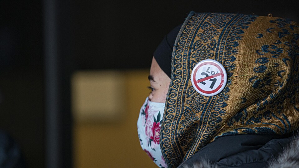 Une femme masquée porte un voile sur lequel est épinglé un macaron contre la loi 21.