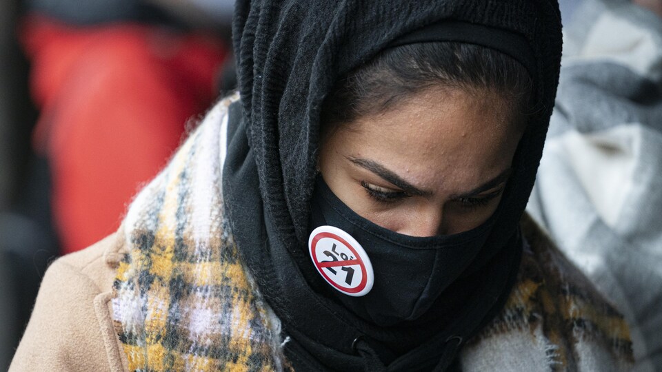 Une femme voilée porte un masque sur lequel se trouve un macaron exprimant son opposition à la Loi sur la laïcité de l'État.
