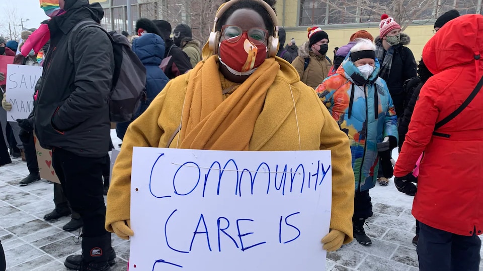 Une personne avec un masque porte une pancarte dans une manifestation sur laquelle on peut lire "Community care is freedom".