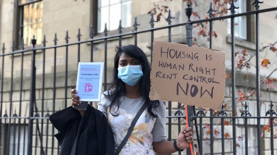 Une femme, le visage couvert d'un masque médical, lors d'une manifestation. Elle tient dans sa main gauche une affiche réclamant le contrôle des loyers, et dans sa main droite un guide pour les locataires.