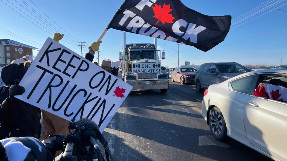 Un camion passe au milieu de la route alors que des gens autour de la route brandissent une affiche et un drapeau.