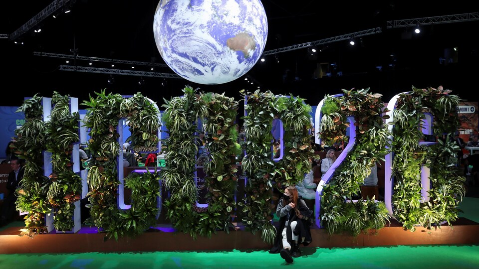 Une femme est assise devant une installation où les lettres du mot COP26 sont composés de plantes. Une représentation de la planète Terre flotte au-dessus des lettres.