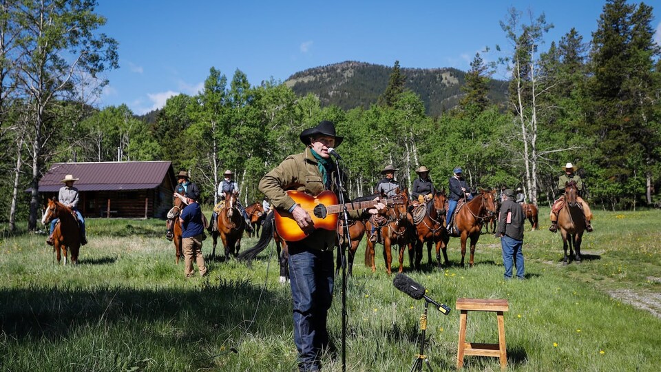 Corb Lund joue de la guitare et chante dans un champ entouré d'arbres. Derrière lui se trouvent des personne sur des chevaux et une montagne.