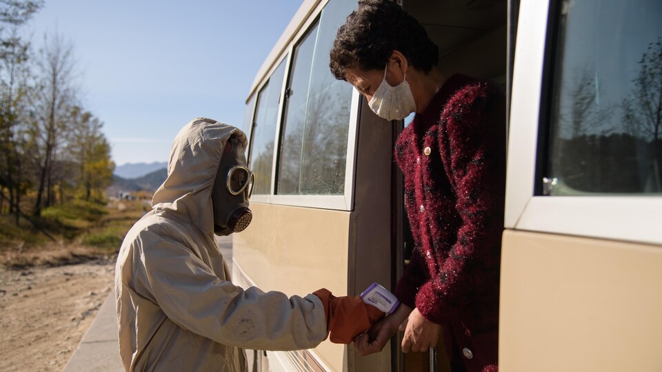 Une personne en combinaison, avec un masque à gaz, prend la température au poignet d'une passagère d'un autobus. 