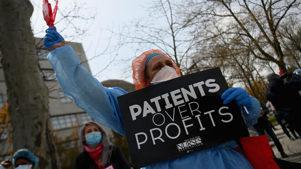 Une personne masquée et vêtue d'une blouse porte une pancarte sur laquelle est écrit : « Patients over profits » (les patients avant les profits).