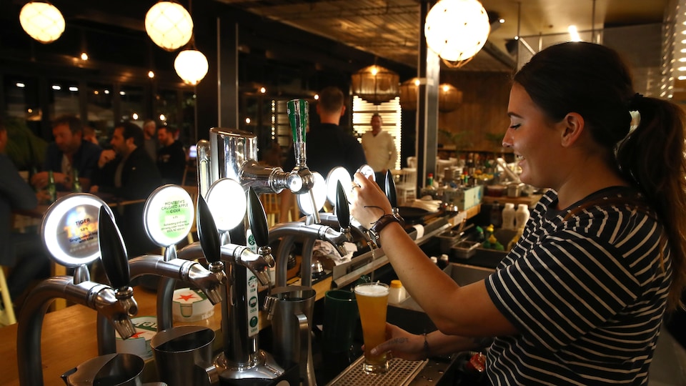 Une femme verse de la bière dans un verre derrière le comptoir d'un bar. Quelques clients sont attablés non loin de là.