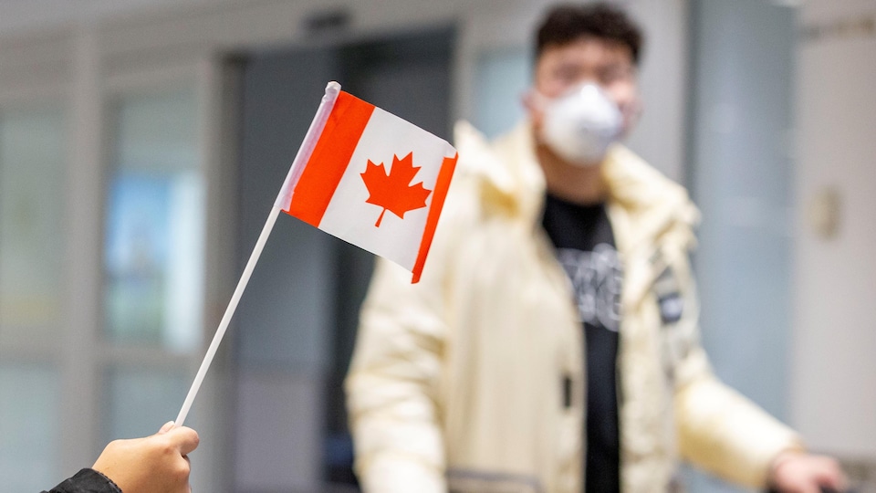 Une main agite un petit drapeau du Canada alors qu'un homme portant un masque marche dans l'aéroport.