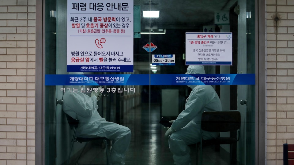 Deux employés d'un hôpital sud-coréen portant des combinaisons stériles discutent assis derrière une porte vitrée.