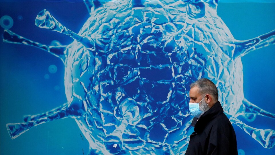Un hombre con una máscara que cubre su rostro camina afuera pegando una imagen muy grande del virus Corona.