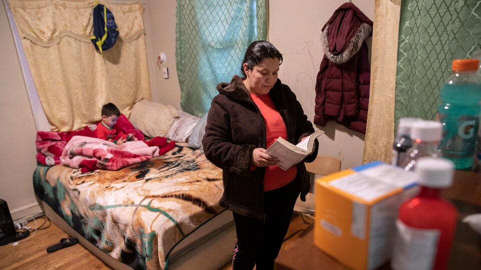 La femme est debout et lit devant un lit où un jeune enfant est assis.