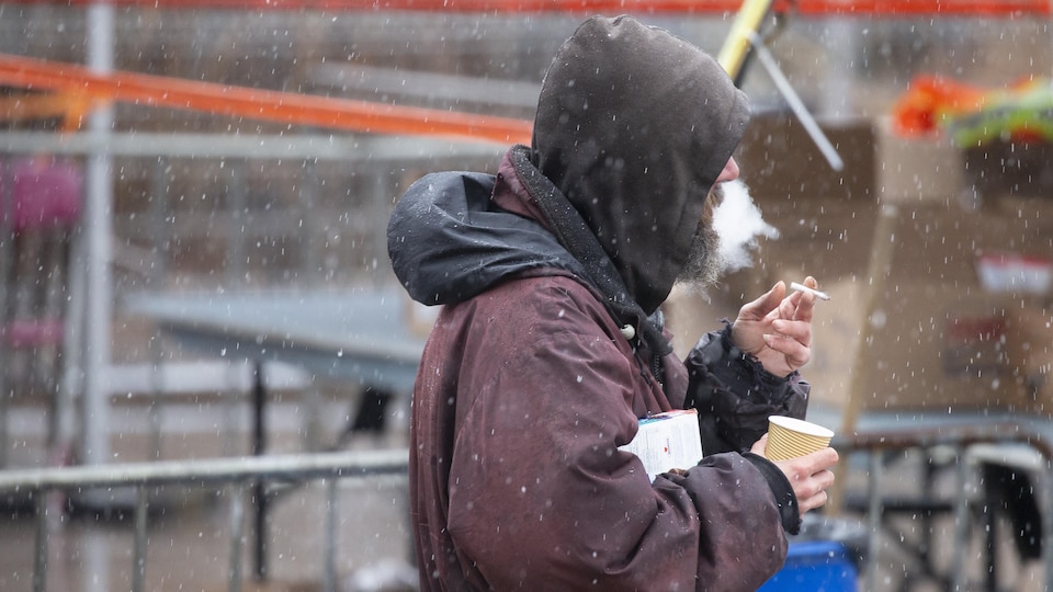Un homme itinérant portant un manteau fume une cigarette sous la neige.