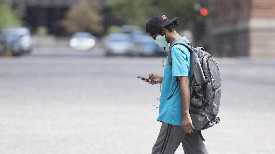 Un homme portant un masque marche dans une rue en regardant son téléphone cellulaire.