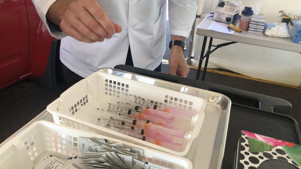 Deux récipients de plastique, l'un rempli de seringues et l'autre de tampons désinfectants, sont placés sur une table devant le pharmacien.