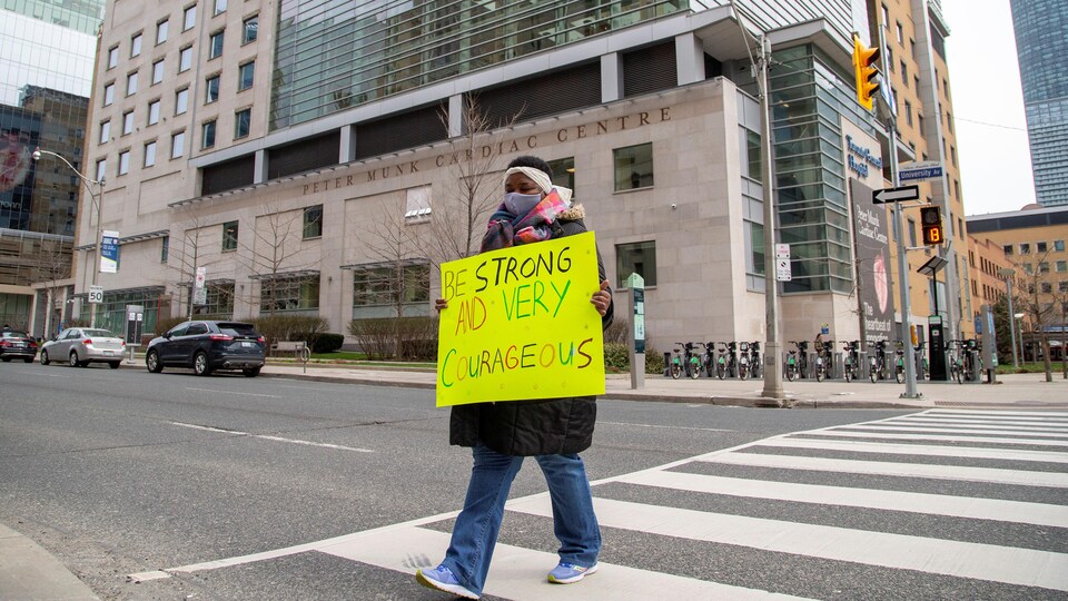 Une femme masquée tient une pancarte devant elle en traversant une rue près d'un hôpital. 