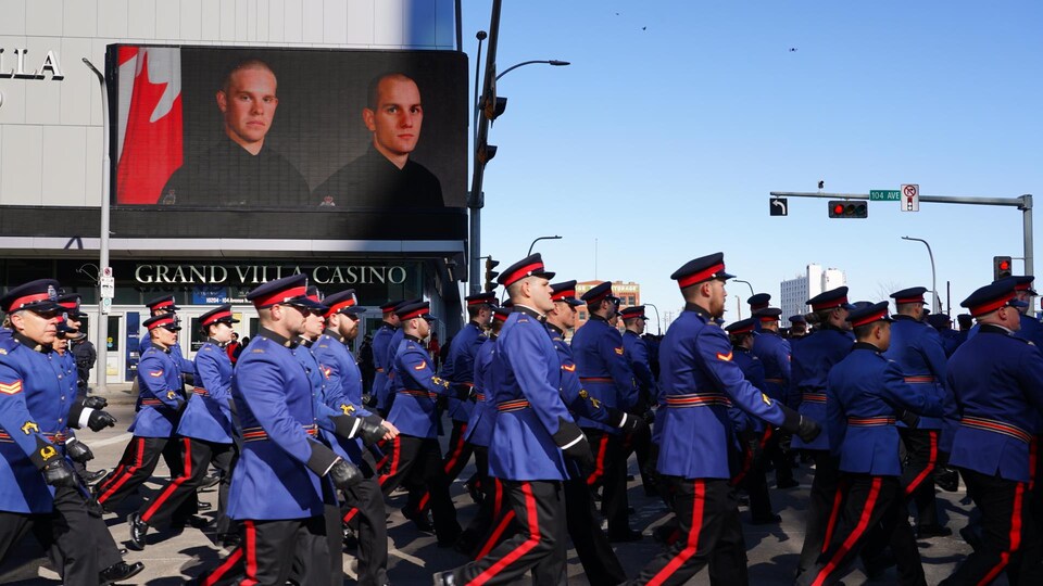 Un cortège de policiers marche au pas devant un immeuble où est affichée la photo de Brett Ryan et Travis Jordan.