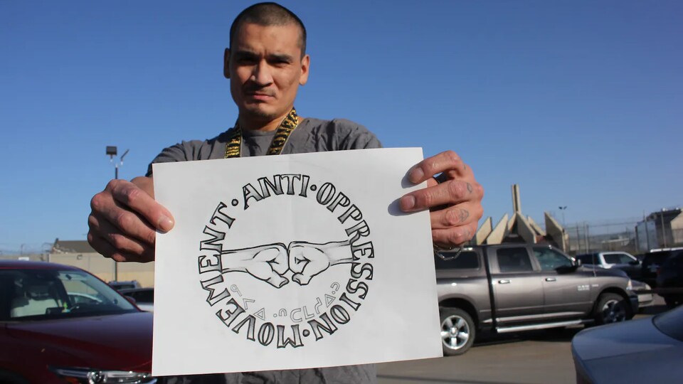 Cory Cardinal montre un dessin qu'il a créé en prison. Il représente deux points qui se touchent avec écrit en rond autour : Mouvement anti-oppression. Il tient ce dessin avec ses deux mots sur un parking.
