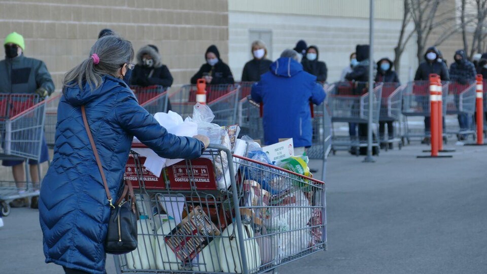 Des gens portant des masques, derrière des paniers d'épicerie vides, font la file à l'extérieur d'un commerce. Au premier plan, une femme vue de dos à la sortie du commerce pousse un panier rempli.