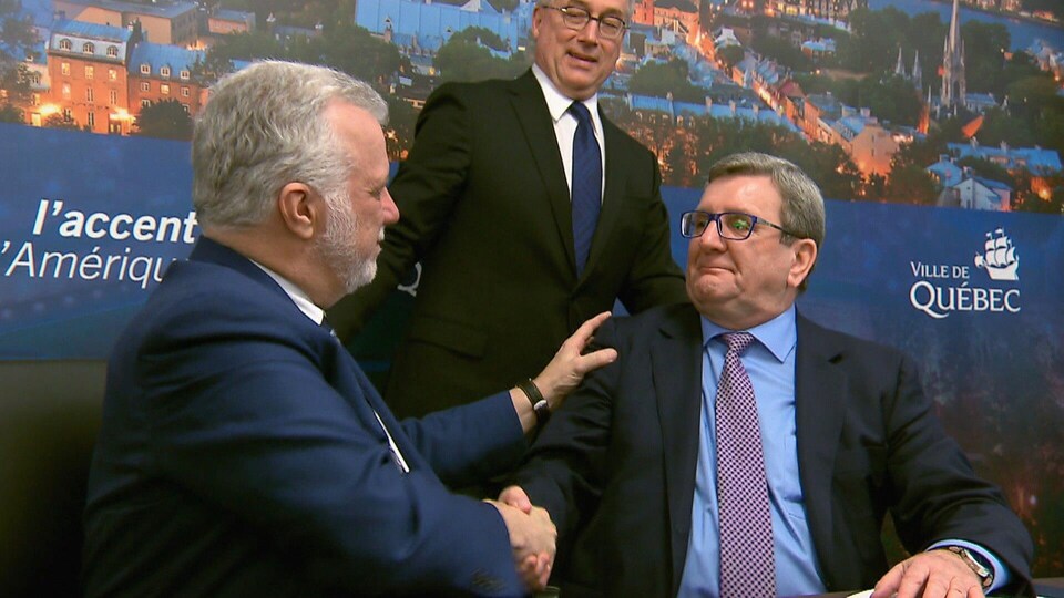 Philippe Couillard et Régis Labeaume se serrent la main sous le regard enjoué de Rémy Normand, alors président du Réseau de transport de la Capitale, lors d'une conférence de presse à l'hôtel de ville de Québec le 15 mars 2018.