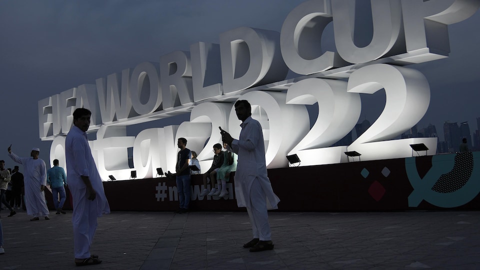 Des hommes posent pour des photos à l'extérieur, devant l'enseigne géante et lumineuse de la Coupe du monde de football 2022.