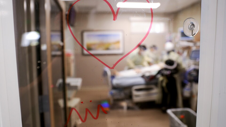 Un cœur est dessiné sur la fenêtre de l’entrée d’une salle d’unité de soins intensifs. On y voit, de façon embrouillée, une personne malade dans un lit d’hôpital qui est entourée d'auxiliaires.