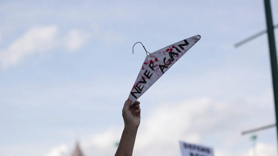 Un manifestant tient un cintre à bout de bras sur lequel on peut lire en anglais : « Jamais plus ».