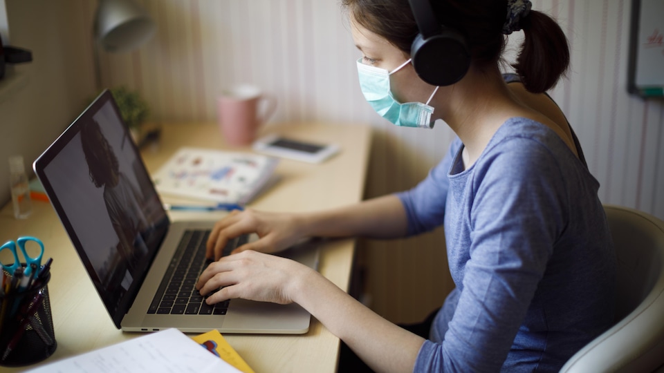 Une adolescente porte un masque de protection au visage et suit sur son ordinateur un cours en ligne à la maison. 