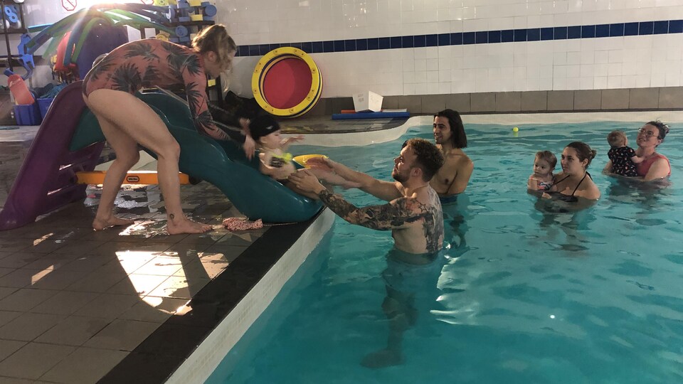 Un jeune enfant glisse sur une glissage qui termine dans une piscine. Des jeunes et des parents se trouvent dans la piscine.