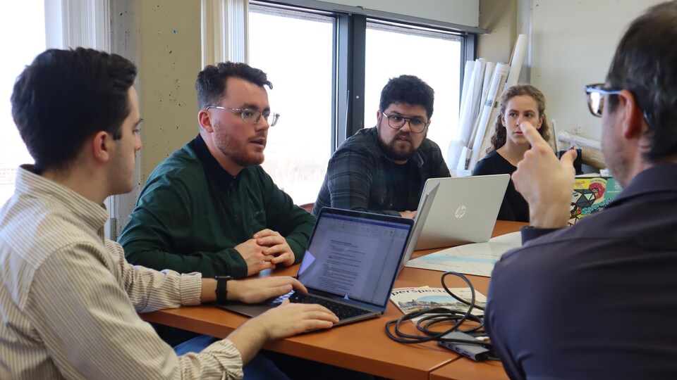 Cinq personnes autour d'une table carrée dans une salle de classe en train de discuter. Certaines ont des ordinateurs portables  ouverts devant elles.