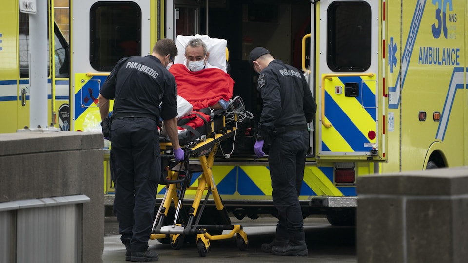 Un homme âgé dans une civière descend d'une ambulance avec l'aide de deux paramédics.