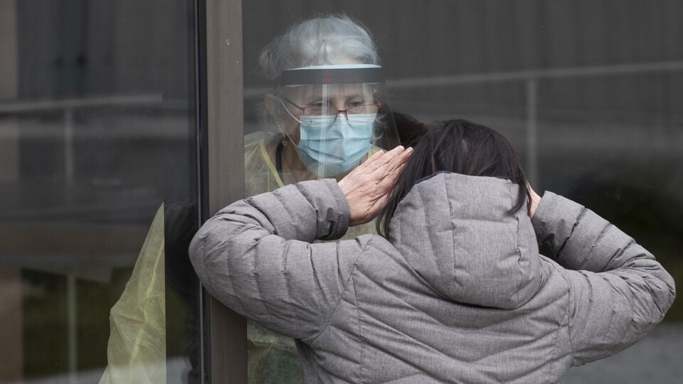 Une femme, de l'autre côté d'une vitre, en communication avec une préposée aux bénéficiaires.