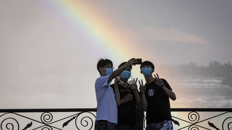Trois personnes portant un masque font le signe de la victoire en se prenant en photo avec un cellulaire devant un arc-en-ciel.
