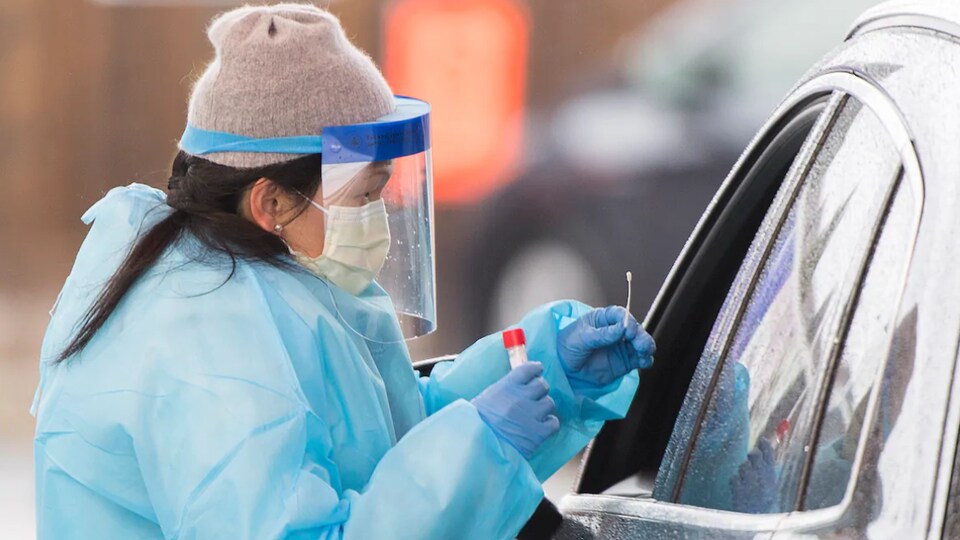 Une femme portant des vêtements et une visière de protection recueille un échantillon de salive d'un automobiliste pour un test de dépistage de la COVID-19.