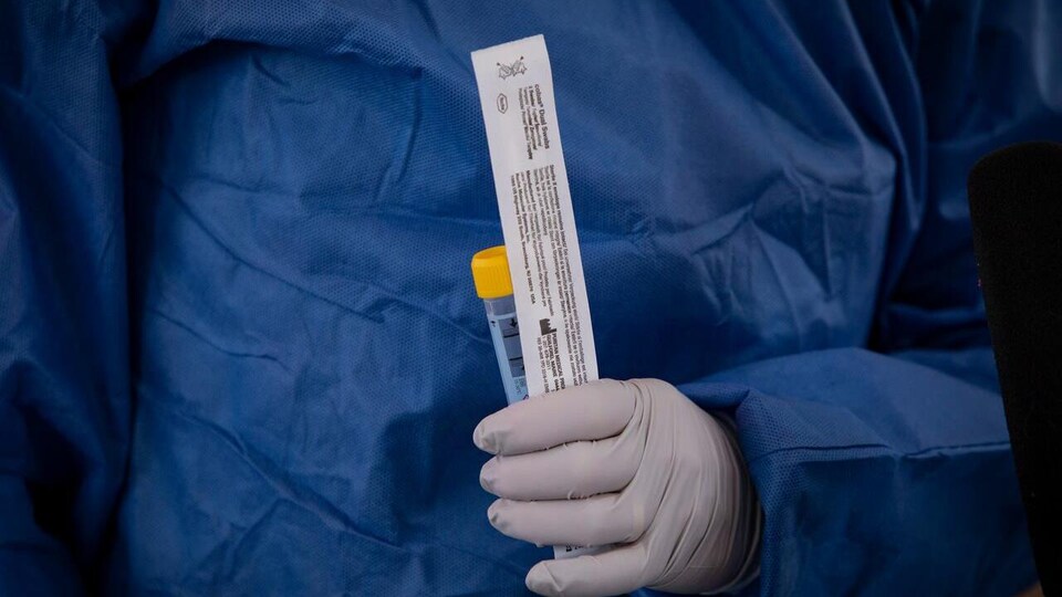 Une main gantée tient un tube et un emballage de plastique dans lequel se trouve un écouvillon servant au dépistage de la COVID-19.