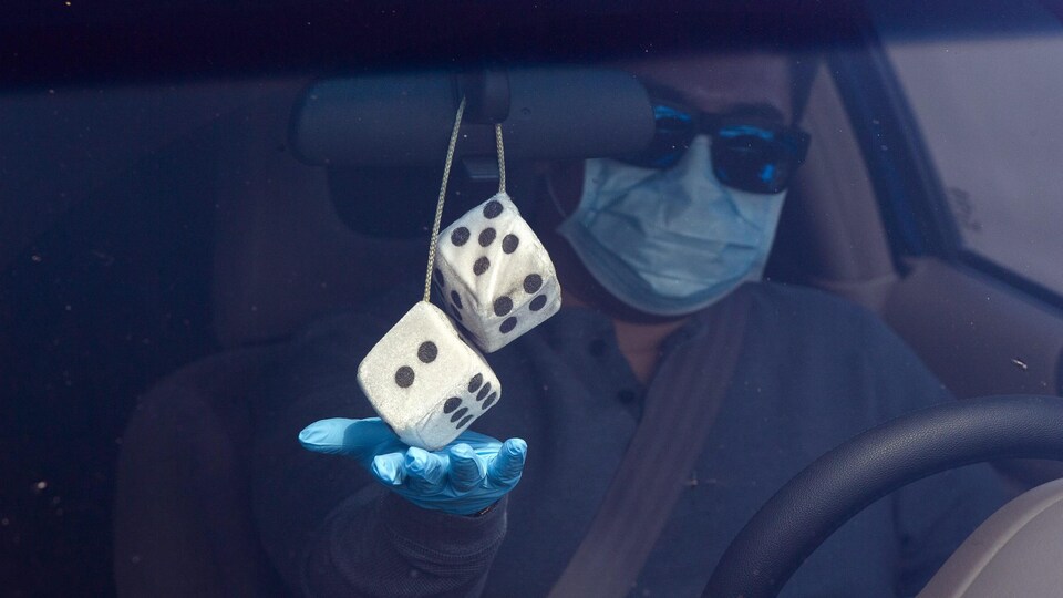 Un homme portant un masque, des lunettes fumées et des gants bleus en latex est dans sa voiture. Il touche les dés en peluche accrochés au rétroviseur intérieur de son véhicule.