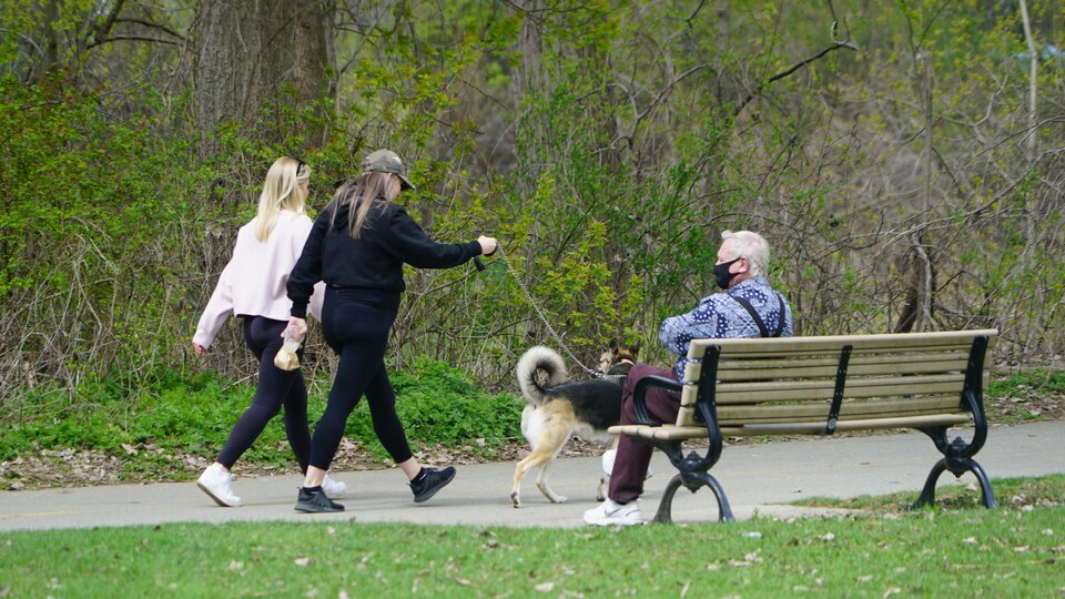Deux femmes faisant marcher un chien passent à côté d'un aîné, assis sur un banc, qui porte un masque.