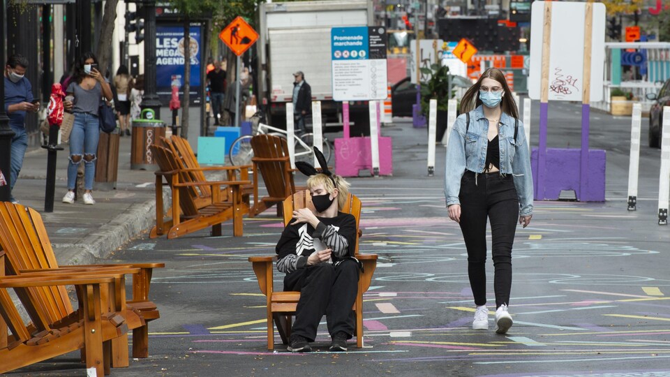 Un homme avec un masque et des oreilles de lapin est assis sur une chaise en plein milieu d'une rue piétonne, dans le Quartier des spectacles, alors qu'une femme, masquée elle aussi, le regarde en marchant.