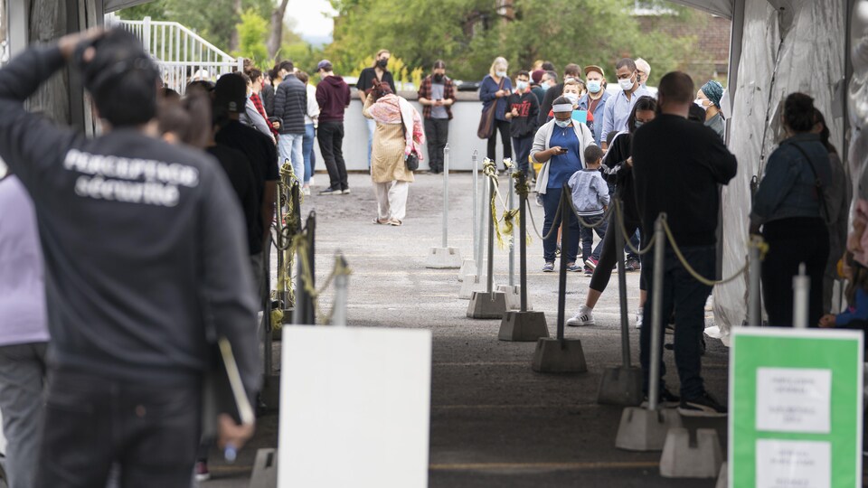 Des dizaines de personnes attendent en file, dehors, sous l'œil d'un agent de sécurité.