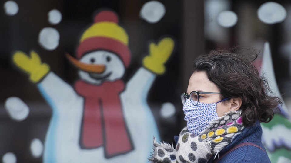 Une femme masquée passe devant une vitrine colorée arborant un dessin de bonhomme de neige.