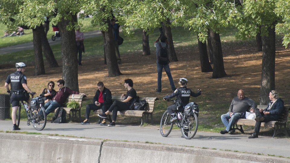 Des Montréalais sont assis sur des bancs par groupes de deux. Les policiers, en vélo, sont arrêtés.