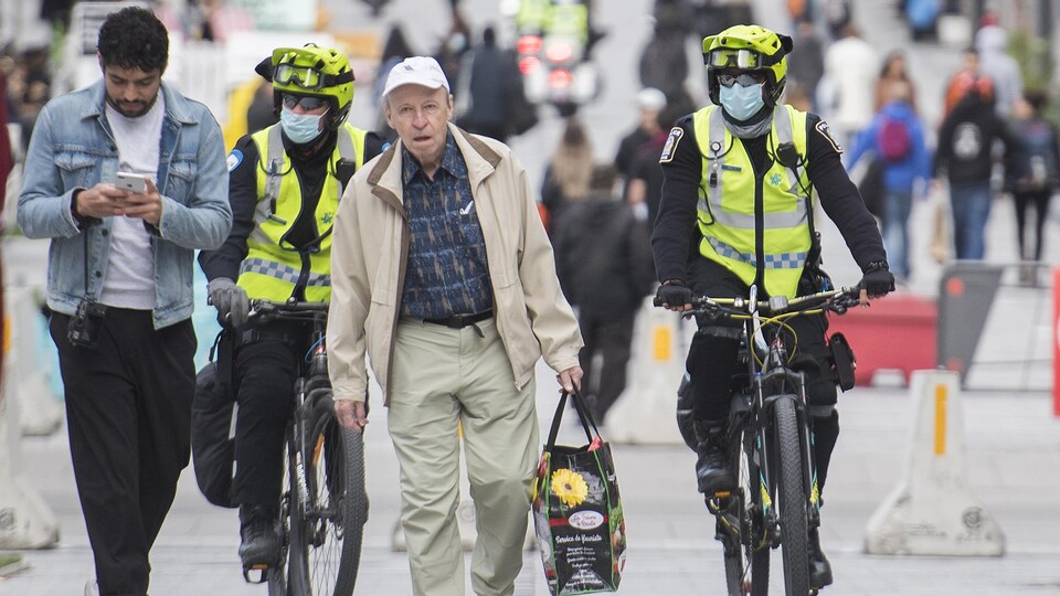 Des policiers en vélo portant des masques se tiennent derrière deux passants qui n'en ont pas.