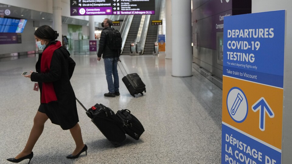 Deux voyageurs à l'aéroport Pearson devant une affiche dirigeant les passagers vers le dépistage contre la COVID-19.