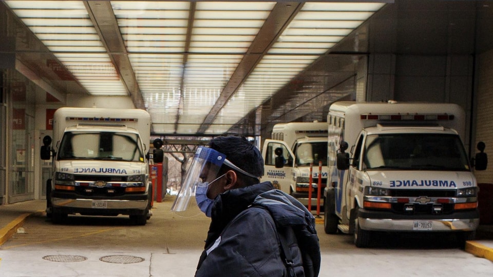 Un travailleur de la santé portant un masque et une visière passe devant des ambulances à l'extérieur d'un hôpital.