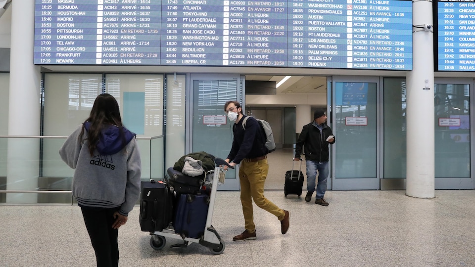 Trois voyageurs dans un aéroport avec des valises, l'un porte un masque.