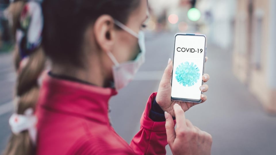 Une femme portant un masque chirurgical regarde son téléphone sur lequel il est écrit COVID-19.