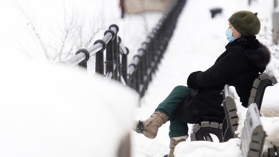 Une femme masquée est assise sur un banc. Elle est entourée de neige.