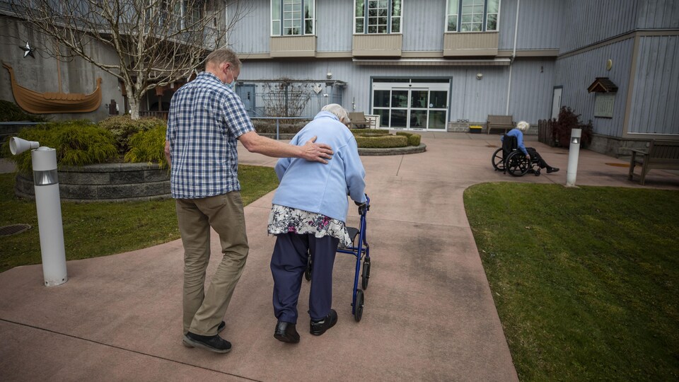 Un homme accompagne sa mère qui marche à l'aide d'un déambulateur à l'extérieur du centre de soins Normanna à Burnaby.